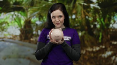 Yeşil parkta elinde domuz kumbarasıyla gönüllü olarak çalışan, mor gömlek giyen, yardım, mutluluk ve toplum desteği taşıyan bir kadın..