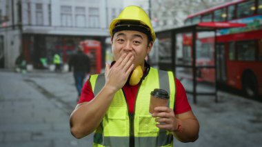 Kasketli ve güvenlik yeleği giymiş bir adam, şehir otobüsü durağında elinde kahve tutarken gülümsüyor. Toplu taşıma ve şehir hayatı belirtileri olan bir arka planda..