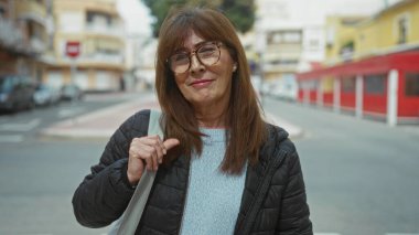 Şehrin sokak ortamında güvenle gülümseyen İspanyol asıllı son sınıf öğrencisi kadın, şehir arka planında, açık havada yakalanmış, açık renk süveter, gözlük ve gündelik ceket, somut modern yaşam tarzı..