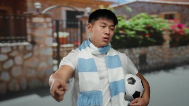 Şehir caddesinde futbol topu tutan genç bir adam uzak bir noktaya doğru hareket ediyor. Canlı şehir ortamında evin yanında eşarp takıyor..