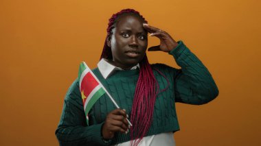 Turuncu arka planda surinam bayrağı olan bir kadın yeşil kazak ve pembe örgüler giyerken düşünceli bir jest yapıyor..