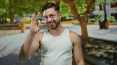 Parkta sakallı bir genç adam yeşil ağaçlar ve rahat bir atmosferle güneşli bir günde beyaz kolsuz bluz giyer ve gülümser..