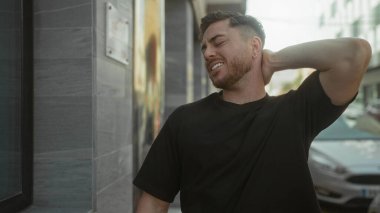 Şehirde sakallı genç bir adam gün ışığında kentsel arka plandan rahatsız olarak boynunu tutuyor, gündüzleri sokakta günlük siyah gömlek giyiyor..