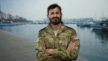Üniformalı bir asker arka planda gemi ve teknelerle dolu hareketli bir limanda, vatansever bir Amerikan bayrağını sergiliyor..