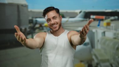 Sakallı genç adam havaalanı terminalinde sevinçle gülümsüyor arka planda kendini uçaklarla kucaklıyor..