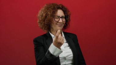 Orta yaşlı, takım elbiseli İspanyol bir kadın, canlı kırmızı bir duvara el hareketi yaparak izole edilmiş bir stüdyoda güven ve iletişimi harmanlıyor..