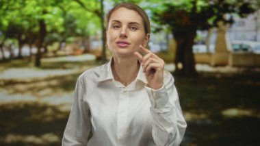 Parkta beyaz gömlekli bir kadın var. Etrafı yeşil ağaçlarla çevrili. Güneşli bir gün.