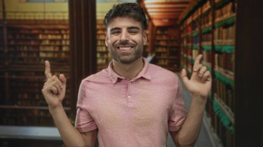 Pembe gömlekli genç adam gülümsüyor ve binadaki ahşap kütüphane rafını işaret ediyor..