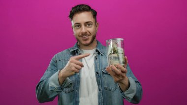 Sakallı genç bir adam, izole edilmiş pembe bir arkaplanı işaret eden bir kavanoz dolusu dolarımızı tutuyor. Kot ceket giyiyor ve kameraya içten bir şekilde gülümsüyor.