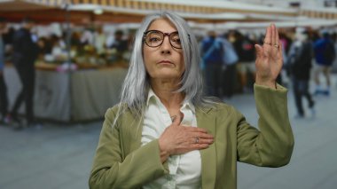 Gri saçlı bir kadın açık hava pazarında küfür jesti yapıyor. Etrafı bulanık müşterilerle çevrili. Güven ve samimiyet duygusu taşıyor..