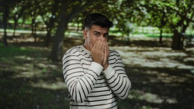 Çizgili tişörtlü genç adam endişeli görünüyor, güneşli bir parkta ağzı kapalı, gür ağaçlarla çevrili, sakin, açık havada derin düşüncelere dalmış bir ruh hali içinde..