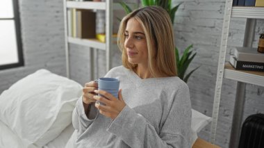 Yatak odasında rahat bir kadın elinde bir kahve fincanı tutuyordu. Etrafı kitaplar ve bitkilerle doluydu. Parlak sabah güneşi iç mekanı aydınlatıyordu..