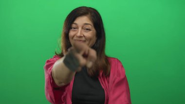 Stüdyoda pembe ceketli, gülümseyen ve kamerayı yeşil ekrana gösteren bir kadın..