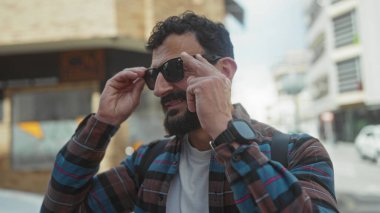Sakallı ve güneş gözlüklü bir adam güneşli bir sokakta dikiliyor. Ekose gömlek ve saat giyiyor. Şehir ortamında kendine güveni tam..