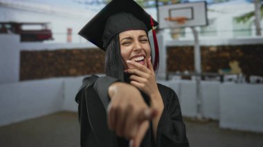 İspanyol kadın mezuniyet şapkası ve cübbesiyle dışarıda basketbol sahasında gülüyor, başarısını neşeyle kutluyor..