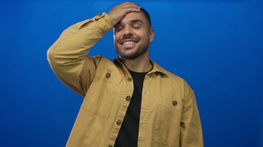 Parlak mavi stüdyoda kahverengi ceketli ve siyah gömlekli bir adam gülüyor ve alnına dokunuyor..