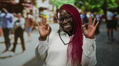 Sokak şehrinde kırmızı örgüler ve gözlüklerle gülümseyen bir kadın güzel bir jest sergiliyor, mutluluğu ifade ediyor, canlı bir atmosferi olan şehir arka planında sıradan bir tarz sergiliyor..