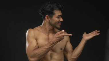 Çıplak göğsü açık avuç içi gösteren, stüdyo ortamında hafifçe gülümseyen, kendine güvenen bir adam..