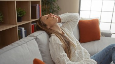 Sarışın kadın şık bir oturma odasında rahat bir kanepede dinleniyor, rahat evinde rahat bir anın tadını çıkarıyor, yumuşak ışıklandırma ve modern dekor aksanıyla..