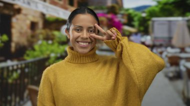 Terasta hardal süveter giymiş bir kadın arka planda bulanık bir açık hava restoranıyla barış işareti sergiliyor. Büyüleyici bir İspanyol atmosferi olduğunu gösteriyor..