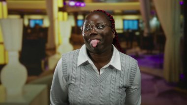 Bir otel ortamında, neşeli bir şekilde dilini dışarı çıkaran bir kadın, canlı ışıklandırma ve modern dekorla, neşeli ve esprili bir atmosfer yayıyor..