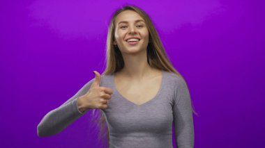 Genç bir kadın, parlak mor bir arkaplan karşısında baş parmağıyla gülümsüyor, gri bir bluz giyiyor, sade ve temiz bir ortamda mutluluk ve pozitifliği iletiyor..