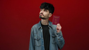 Kırmızı arka planda pasaport taşıyan Türk adam, düşünceli bir ifadeyle uyruğunu ve kimlik bilgilerini sergiliyor.
