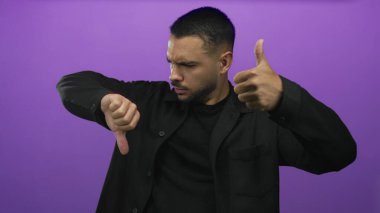 Siyah tişört giyen Latin asıllı genç adam mor stüdyo ortamında baş parmak işareti yapıyor ve kabul etmiyor..