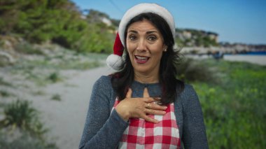 Orta yaşlı İspanyol kadın kırmızı Noel Baba şapkası ve damalı önlük takıyor sahilde el ele tutuşuyor; bayram minnettarlığı.