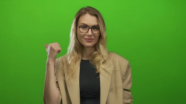Gülümseyen ve baş parmağını geriye doğru gösteren gözlüklü, parlak yeşil arka planda bej ceketli bir kadın..