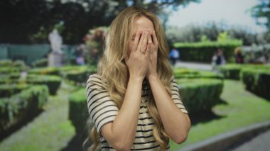 Bir park sahnesinde hayal kırıklığına uğramış bir kadın ellerini kapatıyor, genç sarışın dışarıda yeşil çitlerin arasında görünüyor, bir facepalm jesti ifade ediyor.