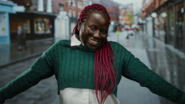 Yağmurlu bir şehir caddesinde kollarını açmış gülümseyen bir kadın şehir ortamında neşesini ve varlığını vurguluyor..