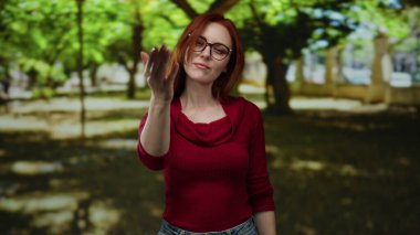 Açık hava parkı ortamında, kırmızı saçlı ve gözlüklü, yemyeşil ağaçlarla çevrili bir kadın, gün ışığında kararlı bir ifade sergiliyor..