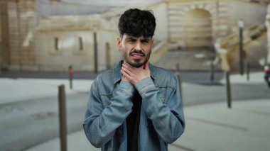 Açık şehir ortamında boğazını tutan kot giyen genç İspanyol bir adam kasabanın arka planına karşı rahatsızlık gösteriyordu..