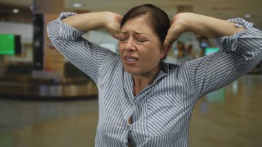 Orta yaşlı mavi çizgili gömlekli kadın havaalanı terminalinde kulaklarını gergin bir ifadeyle kapatıyor..