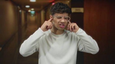 Beyaz gömlekli ve gözlüklü bir adam, loş otel koridorunda parmakları olan kulakları tıkayıp, gözleri kapalı ve ağzı açık bir şekilde rahatsız olur..