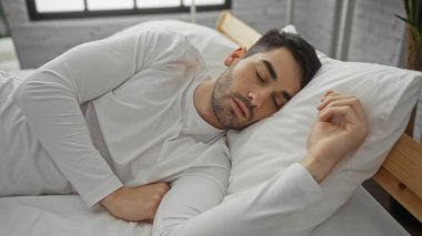 Yakışıklı genç adam modern yatak odasında huzur içinde uyuyor. Beyaz yatak odası ve doğal ışık pencereden içeri akıyor. Huzurlu ve rahatlatıcı bir atmosfer yaratıyor.
