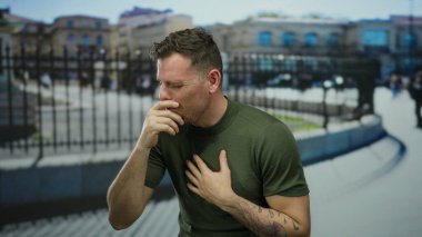 Şehir dışında hapşıran genç bir adam yeşil gömlekli ve dövmeli bir adam. Gün boyunca bulanık şehir arka planına karşı görünür..