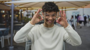 Gözlük takan İspanyol genç adam, iki elini de sokak ortasında şemsiyelerin altında işaret işareti yapıyor..