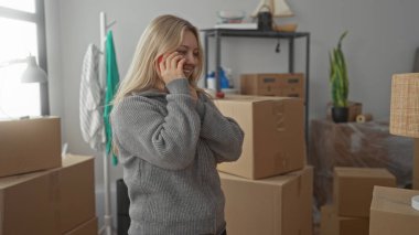 Parlak oturma odasında, paketlenmiş kutularla konuşan, yeni ev için heyecanlanan, rahat gri bir kazak giyen, sarı saçlı, yeni başlangıçların pozitif enerjisini gösteren bir kadın..