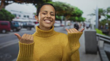 Neşeli bir gülümseme ile 'buraya gel' diyen bir kadın, arka planda bulanık yayalar ve araçlarla dolu kalabalık bir şehir caddesinde rahat sarı bir kazak giyiyor..