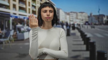 Latin kökenli genç bir kadın açık renk bir bluz giyiyor ve elini çenesine koyuyor. Düşünceli bir bakışla sokakta duruyor..