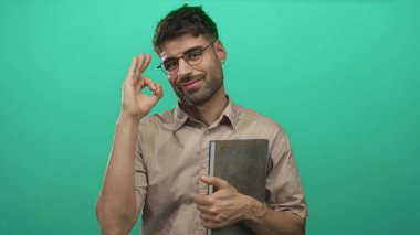 Gözlüklü gülümseyen adam kitabı tutuyor ve parlak yeşil stüdyoda el hareketleriyle kabul ediyor..