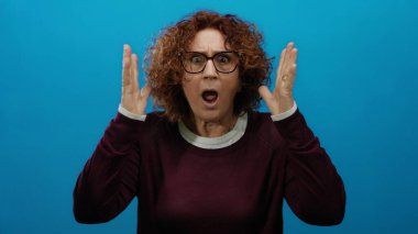 Kıvırcık saçlı, gözlüklü, elleri mavi arka planda, kahverengi saçlı, kahverengi kazak giyen orta yaşlı, duygusal bir kadın.