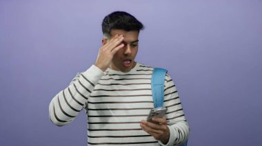 Mor arkaplan önünde telefona şaşırmış genç adam çizgili gömlek ve mavi sırt çantası giyiyor.