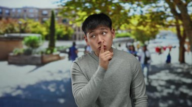 Genç adam, dudağını tutarak canlı bir şehir caddesinde sessizliği işaret ediyor. Arka planda bulanık insanlar ve ağaçlar var..
