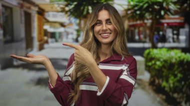 Genç sarışın kadın, şehir caddesinde güneş ışığı altındaki dükkanlarla dolu bir avuç içini işaret ediyor; davet önerisi.