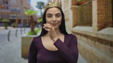 Taçlı bir kadın, güneşli bir caddede güven içinde duruyor, zarafet ve gençlik cazibesini kentin arka planında somutlaştırıyor, kraliyet ve günlük gerçekliğin bir karışımını öneriyor..