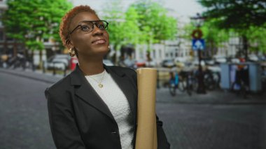 Siyah ceketli kadın, şehir caddesinde yeşil yeşilliklerin altında bina cephesi ve hırsla planları tutuyor..