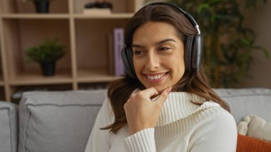 Oturma odasındaki koltukta oturmuş kulaklık takan kadın gülümsüyor. Arka planda raflar, rahat bir iç mekan izlenimi uyandırıyor..